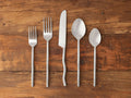 Oaklynn Five Piece Place Setting: two matte silver stainless steel forks, knife, and two spoons laid vertically on a wooden surface.