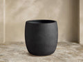 Textured, dark gray Ojai Outdoor Planter, barrel-shaped, on a light stone patio against a beige wall.