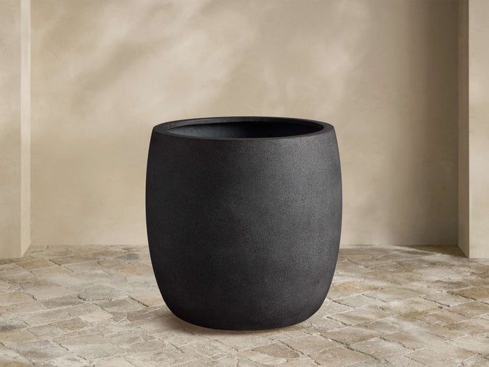 Textured, dark gray Ojai Outdoor Planter, barrel-shaped, on a light stone patio against a beige wall.