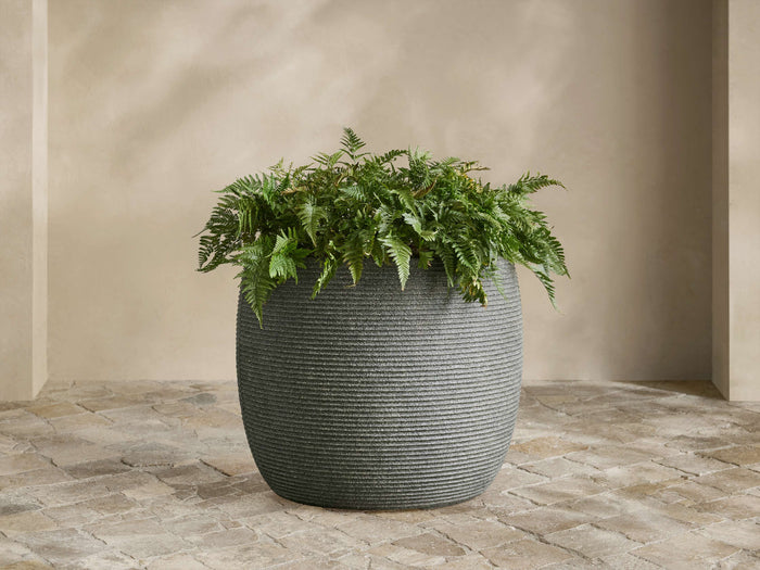 Dark gray Ojai Ribbed Outdoor Planter, rounded with horizontal ridges, containing a lush green fern, on stone pavers.