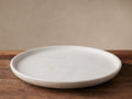 Round Omo Marble Platter, white with natural beige veining, angled on a dark wooden table against a beige wall.