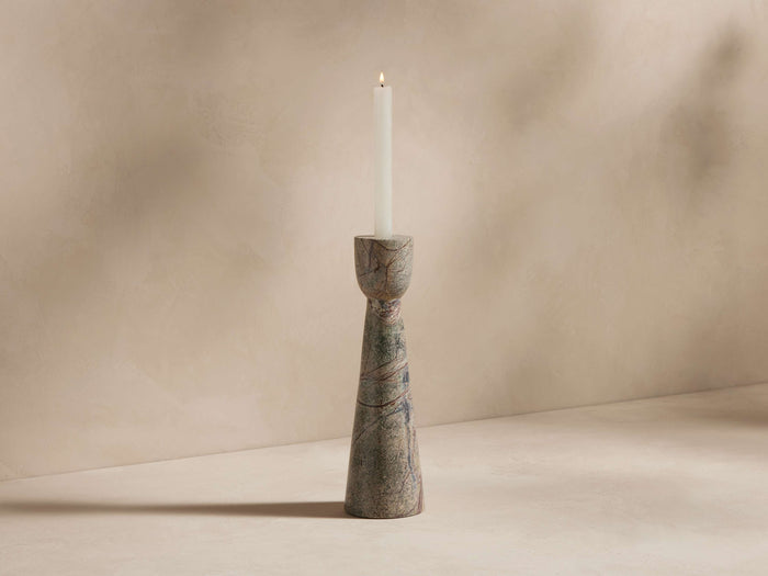 A tall, slender marble candle holder with a smooth, swirling pattern, holding a single lit white taper candle against a neutral background.
