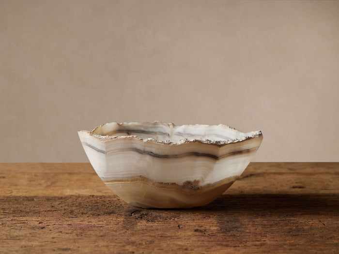 Onyx Bowl with a wavy, organic rim, showing natural cream, brown, and gray bands, sitting on a rustic wooden table.