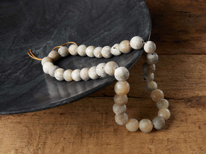 A strand of Onyx Variegated Beads with cream and gray tones, resting on a dark bowl, displayed on wooden surface.