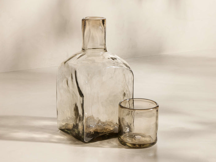 Front view of the Organic Carafe Set, with a tall, square-based carafe and a short glass, both crafted from subtly tinted, textured recycled glass.