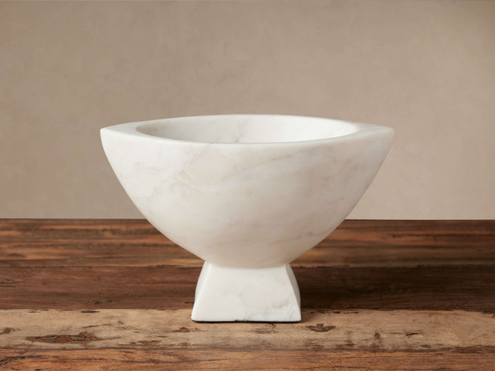 White marble Oslo Footed Bowl, hand-carved, with a flared top and solid rectangular foot, displaying natural veining on a wooden surface.