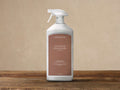White Arhaus Outdoor Upholstery Cleaner spray bottle with brown label, on a wooden table against a beige wall.