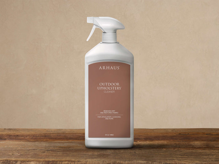 White Arhaus Outdoor Upholstery Cleaner spray bottle with brown label, on a wooden table against a beige wall.