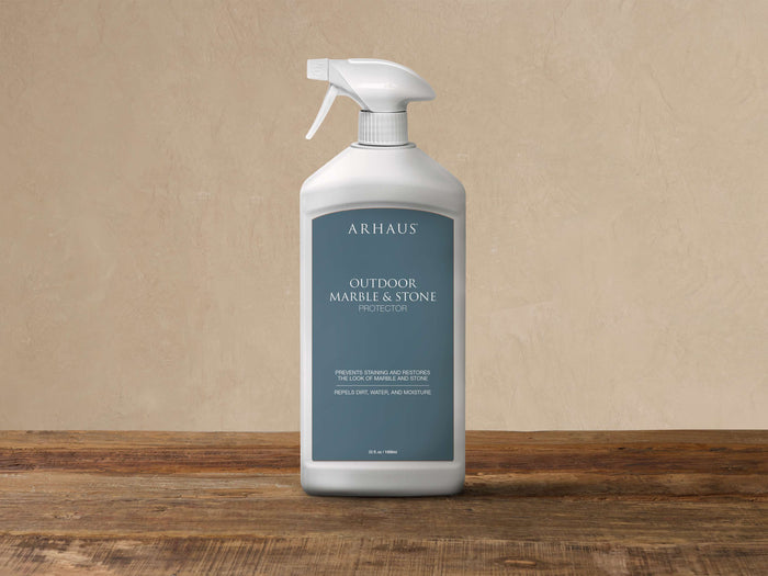 White ARHAUS Outdoor Marble & Stone Protector spray bottle with dark teal label, on a wooden surface against a light wall.