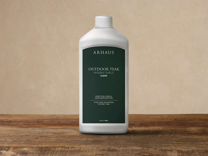 White plastic bottle of Arhaus Outdoor Teak Invisible Shield, 33.8 oz, with a dark green label, standing on wood.