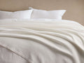 White Paden Linen Ribbed Bed Blanket draped softly on a bed, showing its ribbed texture, with white pillows and a beige wall.