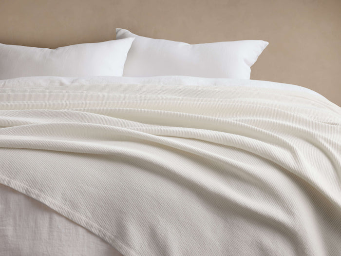 White Paden Linen Ribbed Bed Blanket draped softly on a bed, showing its ribbed texture, with white pillows and a beige wall.