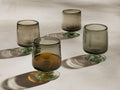 Four hazy gray handblown Paloma double old-fashioned glasses with green-tinted bases, one filled with amber liquid, casting shadows.