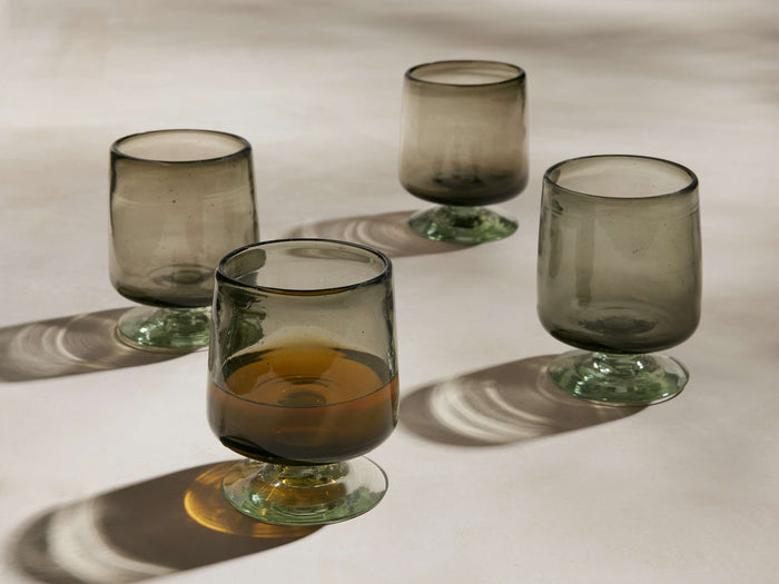Four hazy gray handblown Paloma double old-fashioned glasses with green-tinted bases, one filled with amber liquid, casting shadows.