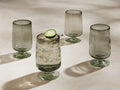 Set of 4 smoky grey handblown Paloma Highball Glasses with footed bases. One filled with ice, liquid, cucumber. Three are empty.