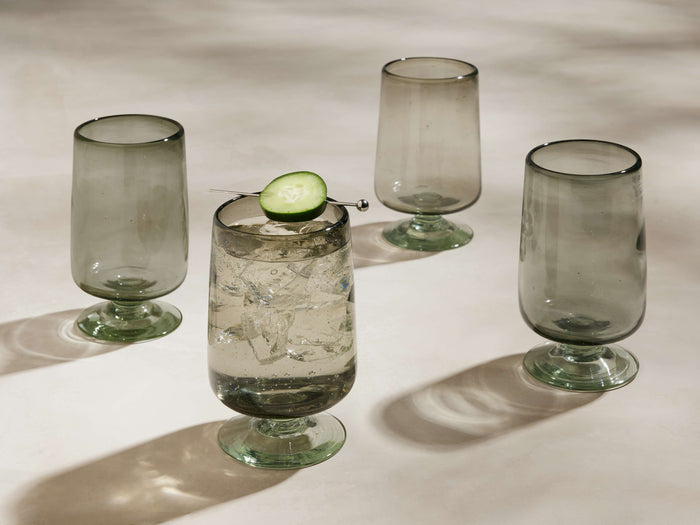 Set of 4 smoky grey handblown Paloma Highball Glasses with footed bases. One filled with ice, liquid, cucumber. Three are empty.