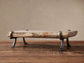 Petrified Wood Serving Board, light-toned with organic edges, held by dark gray wrought iron legs, sitting on a rustic wooden table.