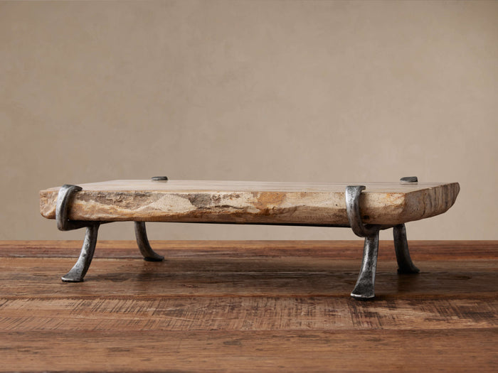 Petrified Wood Serving Board, light-toned with organic edges, held by dark gray wrought iron legs, sitting on a rustic wooden table.