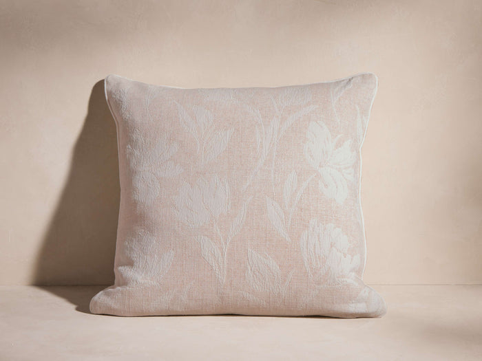 Light pink Phare Pillow with woven off-white floral pattern & corded edge, standing upright against a textured background.