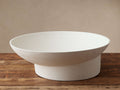White stucco Planes Footed Bowl, wide and shallow, with a textured surface, sits on a rustic wooden table.