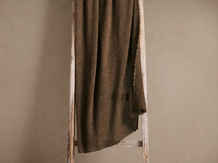 A dark brown textured blanket draped over a rustic wooden ladder against a plain beige background.
