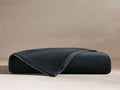 A neatly folded dark grey terry cloth towel with satin trim on a neutral surface, showcasing its plush texture.