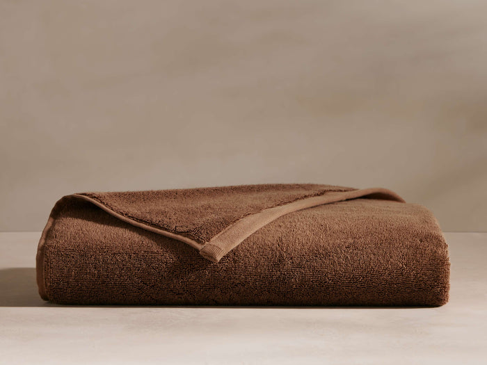 A neatly folded brown towel with a visible textured surface, placed on a light surface against a neutral background.