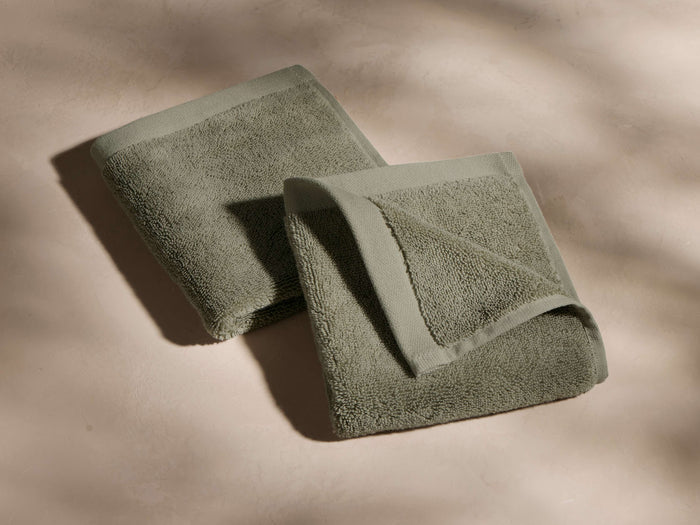 Two neatly folded gray-green towels with visible texture, placed on a smooth, light beige surface with soft shadows.