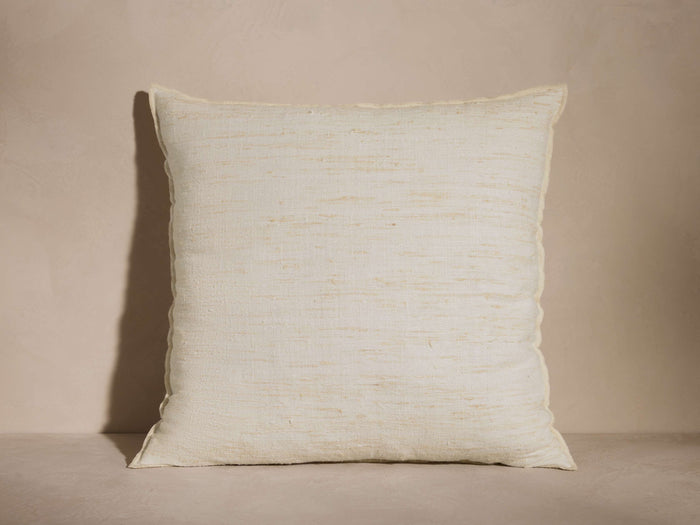 Square off-white Raw Silk Pillow with visible hand-spun texture and subtle tan variations, resting upright against a neutral wall.