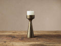 Ren Pillar Holder, conical bronze-finished aluminum, with a lit white pillar candle on a rustic wood surface.
