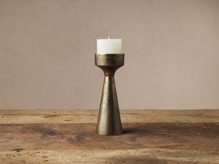 Ren Pillar Holder, conical bronze-finished aluminum, with a lit white pillar candle on a rustic wood surface.