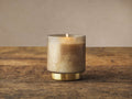 Lit Retreat Vanilla Sugarcane Candle in frosted glass with antique brass base, on a distressed wood table against a beige wall.