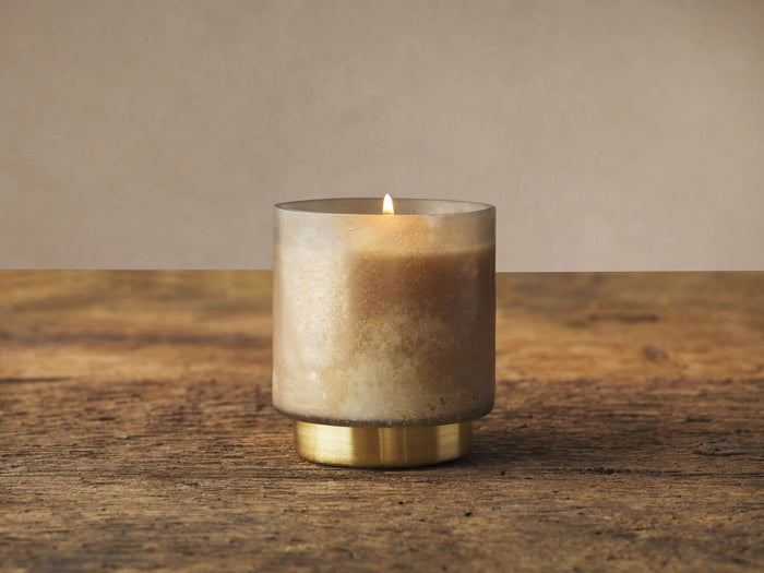 Lit Retreat Vanilla Sugarcane Candle in frosted glass with antique brass base, on a distressed wood table against a beige wall.