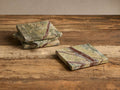 Four Rhodes Coasters, green marble with reddish-brown veins. Three stacked left, one flat right on a rustic wooden surface.