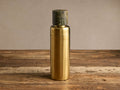 A Rhodes Cocktail Shaker, gold stainless steel, topped with a dark green natural marble cap, standing on a rustic wooden table.