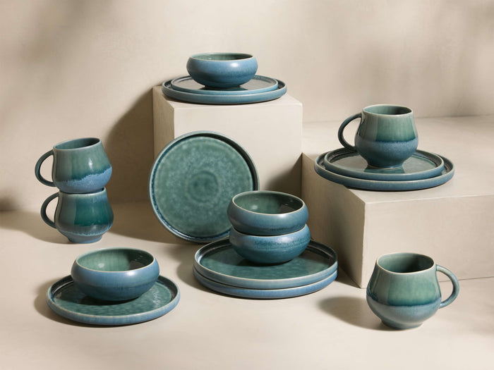 Full view of the blue-green reactive glaze Rhoswen Dinnerware with Cereal Bowls (Set of 16), displayed on neutral blocks.