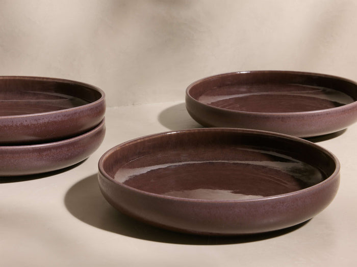 Angled view of four deep plum Rhoswen Pasta Bowls, two stacked, featuring a reactive glaze finish and wide, shallow design.