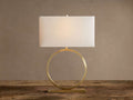 Rhys Table Lamp: Illuminated rectangular light fabric shade atop a gold circular hoop base on a rustic wood surface.