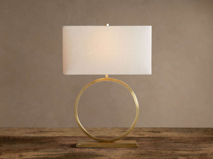 Rhys Table Lamp: Illuminated rectangular light fabric shade atop a gold circular hoop base on a rustic wood surface.