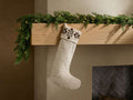 Front view of natural Riad Stocking with dark brown geometric embroidery on cuff, hanging from a garland-decorated mantelpiece.