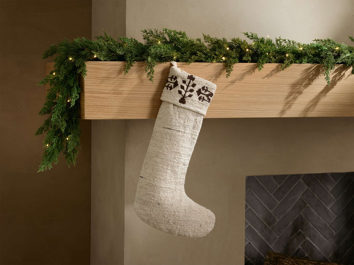 Front view of natural Riad Stocking with dark brown geometric embroidery on cuff, hanging from a garland-decorated mantelpiece.