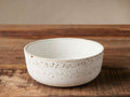Ronan Cereal Bowl, white stoneware with reactive glaze and brown speckles, angled on a rustic wooden table.