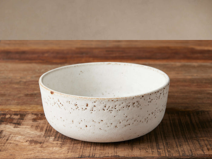 Ronan Cereal Bowl, white stoneware with reactive glaze and brown speckles, angled on a rustic wooden table.