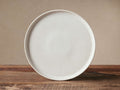 Ronan Dinner Plate, warm ivory reactive glaze stoneware, standing upright on a rustic wood surface against a neutral wall.