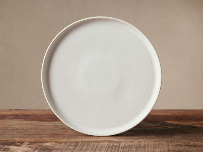 Ronan Dinner Plate, warm ivory reactive glaze stoneware, standing upright on a rustic wood surface against a neutral wall.