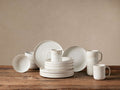 Ronan 16-pc white stoneware dinnerware set, including plates, bowls, and mugs, with speckled reactive glaze on a wooden table.