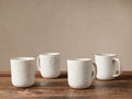 Four Ronan Mugs, ivory stoneware with reactive glaze & brown speckles, on a wood surface against a neutral wall.