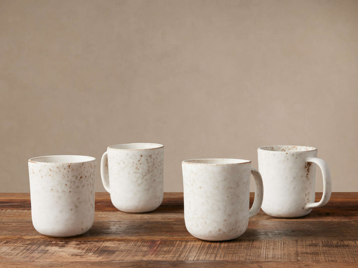 Four Ronan Mugs, ivory stoneware with reactive glaze & brown speckles, on a wood surface against a neutral wall.