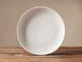 Round Ronan salad bowl, warm ivory stoneware with subtle speckled reactive glaze, on a rustic wooden table.