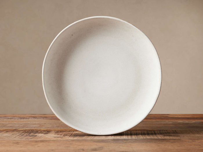 Round Ronan salad bowl, warm ivory stoneware with subtle speckled reactive glaze, on a rustic wooden table.